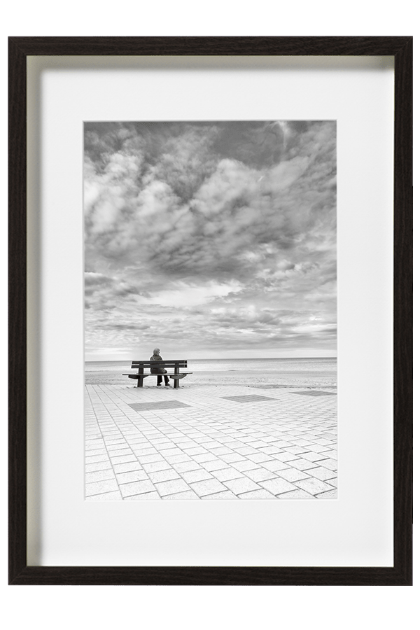 En Belgique a Knokke-Le-Zoute une femme est assise sur un banc face a la mer du Nord. Un ciel très dessiné et un trottoir bien aligné servent ce décor.