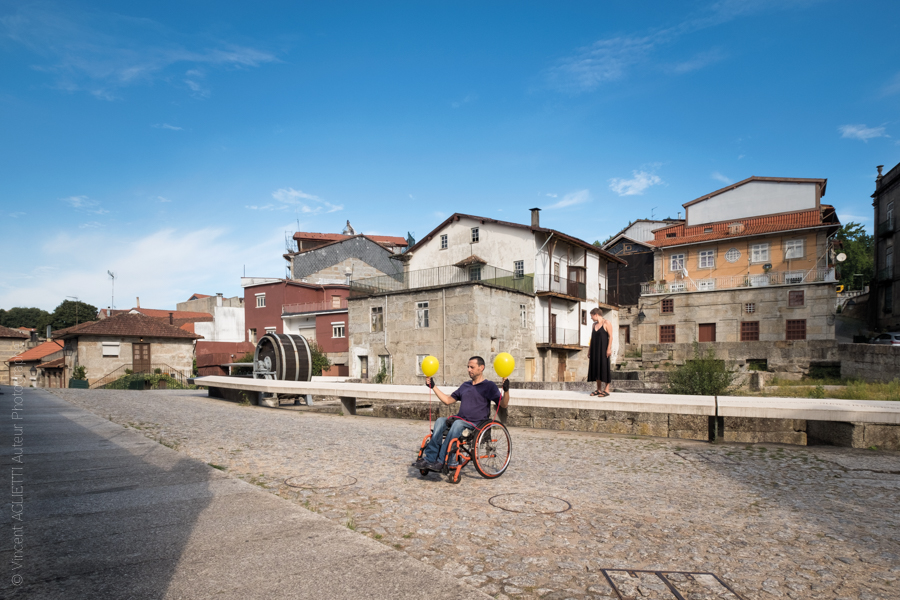 Un homme dans un fauteuil roulant tient deux ballons jaunes, une femme se tient derrière lui.Ils posent Largo do Cidade pour l'exposition Guimaraes, Aqui Nasceu Portugal de Vincent Aglietti et Vincenzo Cirillo.