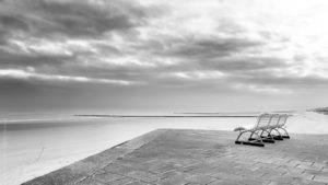 Sans Vis à Vis. Sur les bords de la plage d'Ostende. Un banc vide surplombe la Plage, des traces de pas filent vers la mer. Le ciel est menaçant.