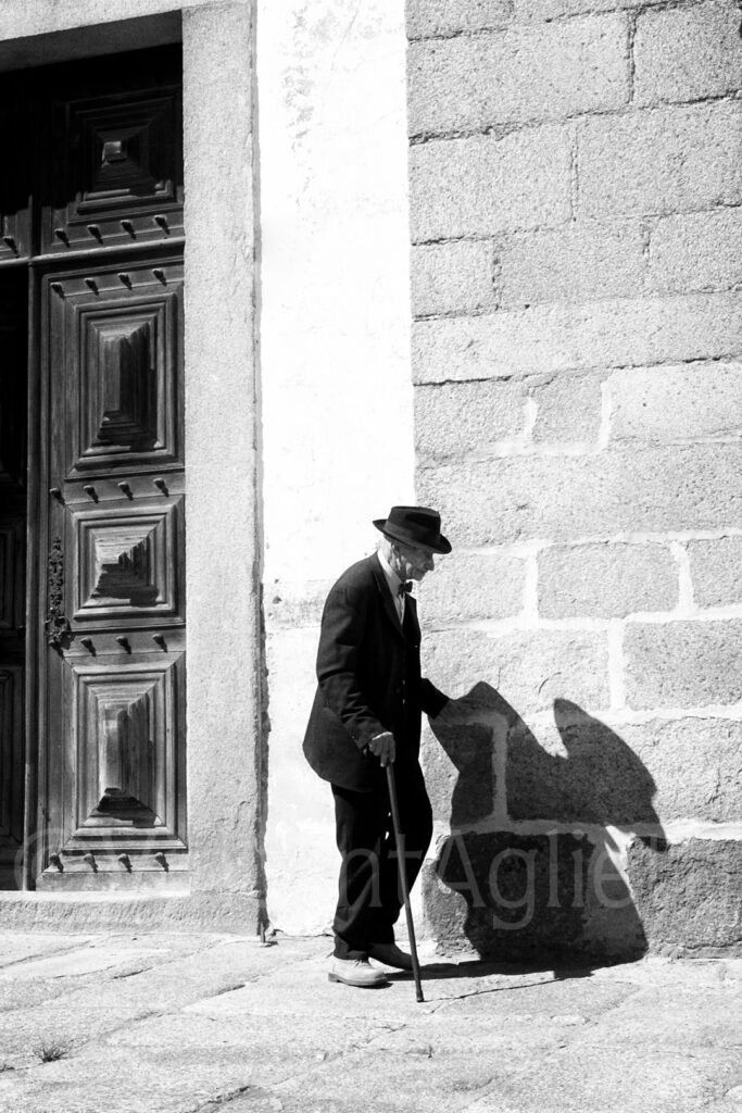 Vieil homme marchant avec une canne dans une rue d'Évora, son ombre se projette sur un mur de pierre.