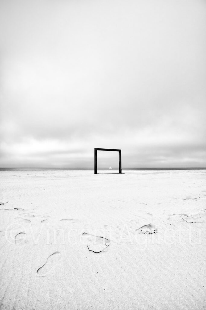 Voilier au loin cadré par une douche de plage à Knokke-le-Zoute, photographie en noir et blanc