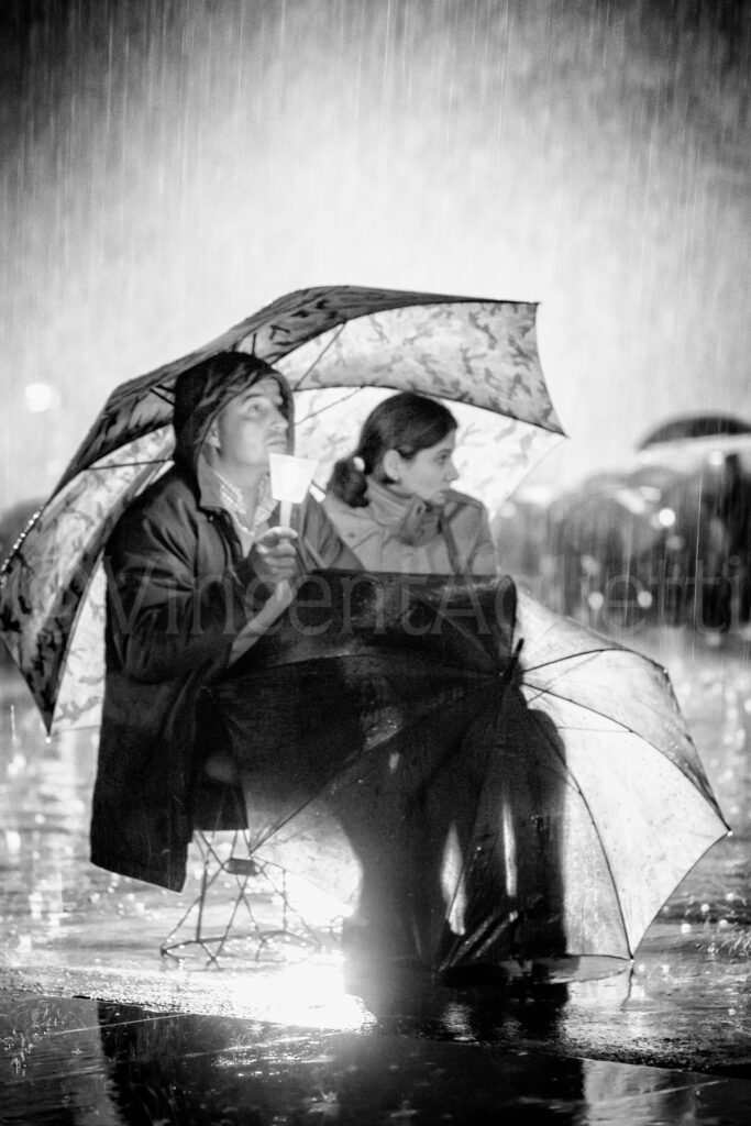 Couple de pèlerins assis sous un parapluie pendant une pluie battante sur l'esplanade du sanctuaire de Fátima.